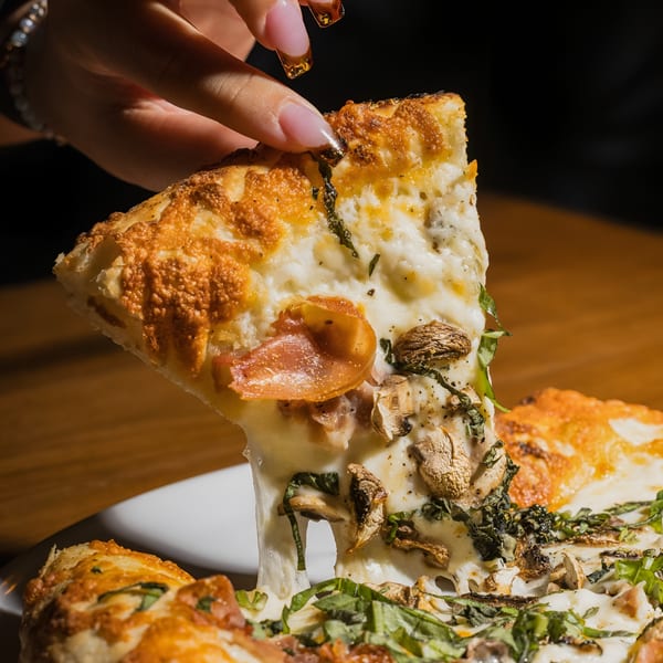 A hand lifts a slice of pizza with melted cheese, mushrooms, prosciutto, and basil.
