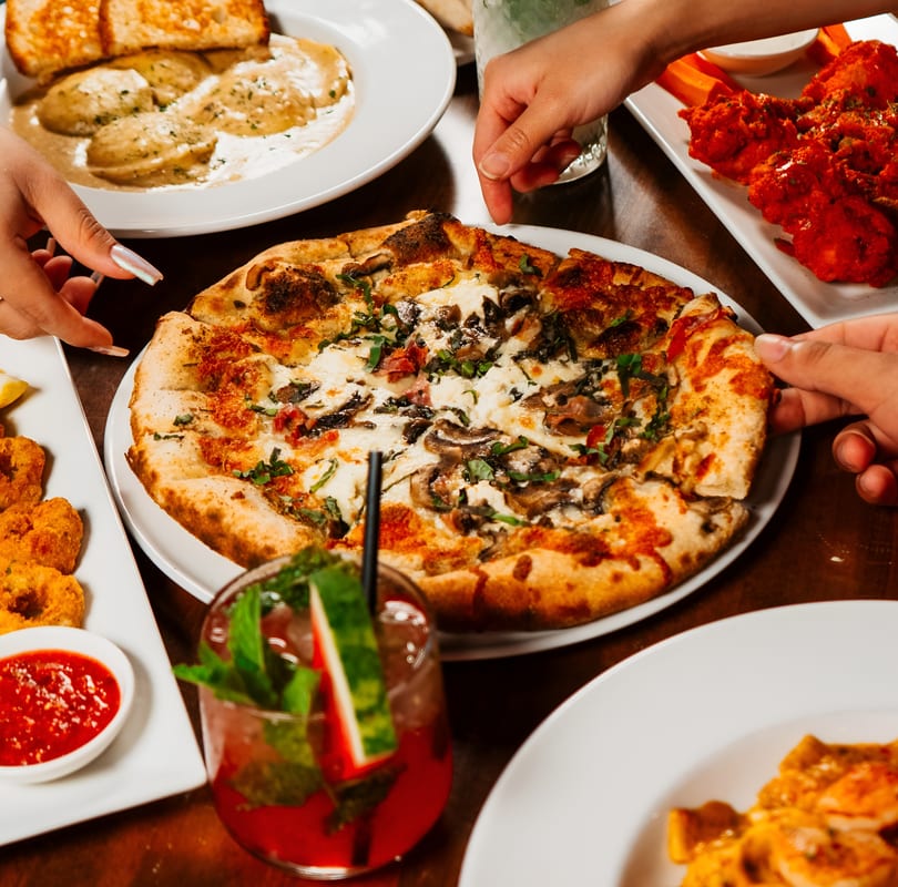Hands reaching for slices of mushroom pizza, surrounded by pasta, wings, and a cocktail.
