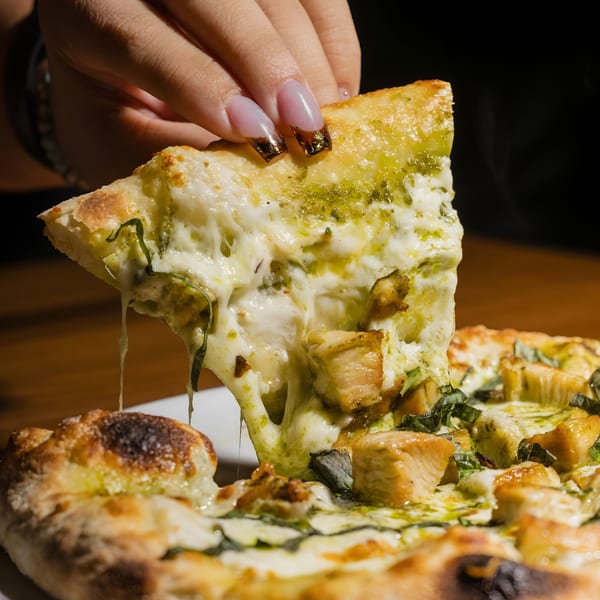 A hand lifts a slice of pesto chicken pizza, with cheese and pesto stretching from the pie.