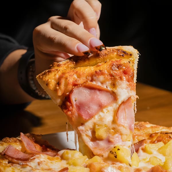 A hand lifts a slice of Hawaiian pizza with melted cheese, ham, and pineapple.